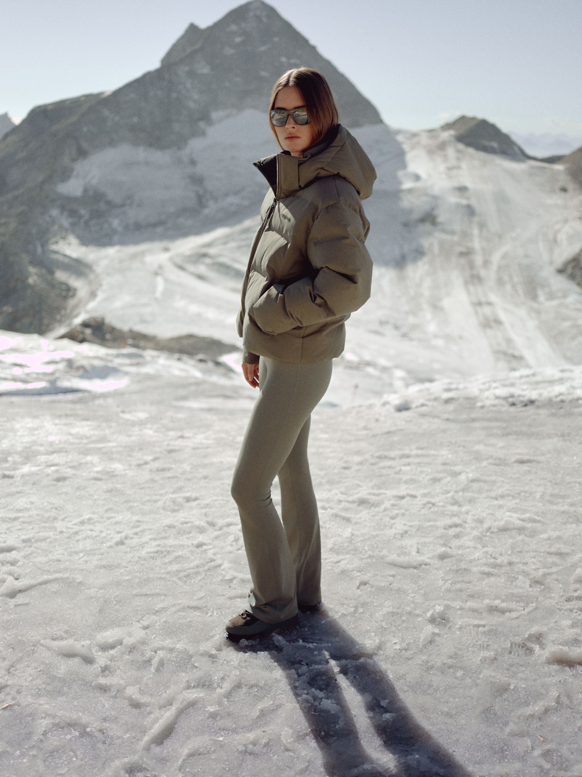 Woman wearing a khaki CASPARA Aurora puffer jacket against a snowy mountain backdrop.